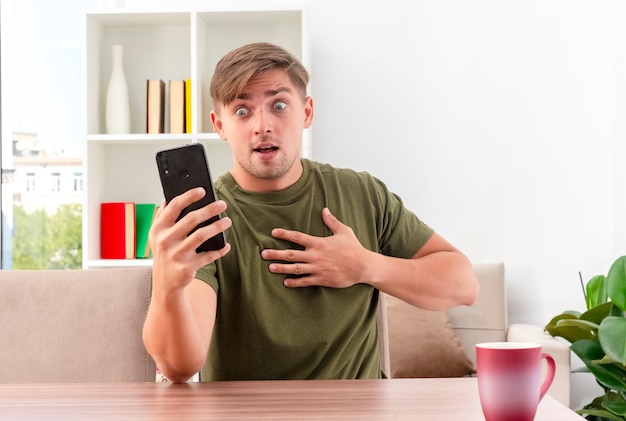 Überraschtes junges blondes hübsches Mann sitzt am Tisch mit Tasse, die Hand auf Brust setzt und Telefon im Wohnzimmer hält