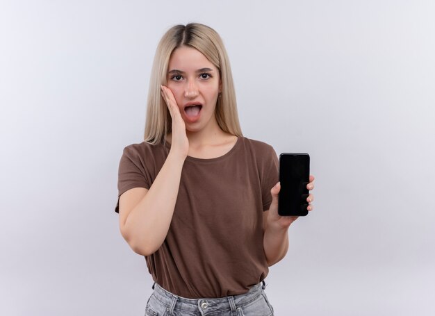 Überraschtes junges blondes Mädchen, das Handy mit Hand auf Wange lokalisierte weiße Wand mit Kopienraum hält