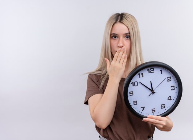 Überraschtes junges blondes Mädchen, das Uhr mit Hand auf Mund auf isolierter weißer Wand mit Kopienraum hält