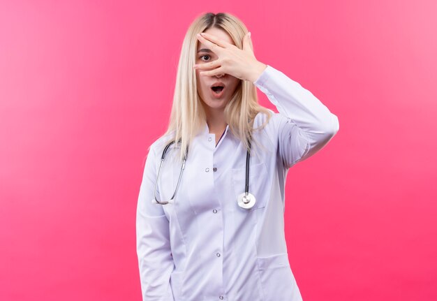 Überraschtes junges blondes Mädchen des Arztes, das Stethoskop im medizinischen Kleid trägt, bedeckte Auge auf lokalisiertem rosa Hintergrund