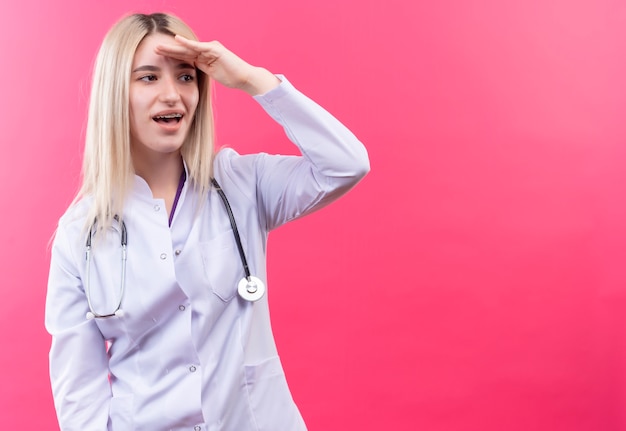 Überraschtes junges blondes Mädchen des Arztes, das Stethoskop im medizinischen Kleid trägt und Entfernung mit Hand auf lokalisiertem rosa Hintergrund schaut