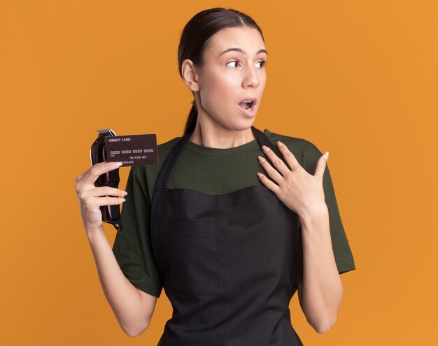 Überraschtes junges brünettes Friseurmädchen in der Uniform hält Haarschneidemaschinen und Kreditkarte legt Hand auf Brust, die Seite auf Orange betrachtet