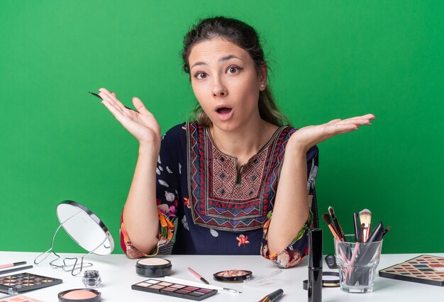 Überraschtes junges brünettes Mädchen, das am Tisch mit Make-up-Tools sitzt und die Hände offen hält, hält Eyeliner