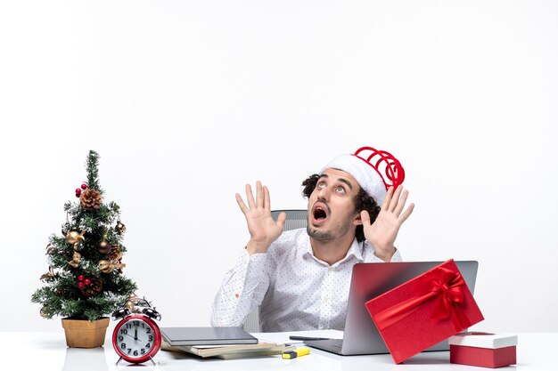 Überraschtes junges Geschäftsmann mit lustigem Weihnachtsmannhut, der über denkendes und feierndes Weihnachten im Büro auf weißem Hintergrund schaut