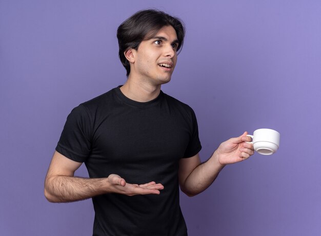 Überraschtes junges gutaussehendes Kerl, das schwarzes T-Shirt hält und Punkte mit Hand an Tasse Kaffee lokalisiert auf lila Wand trägt