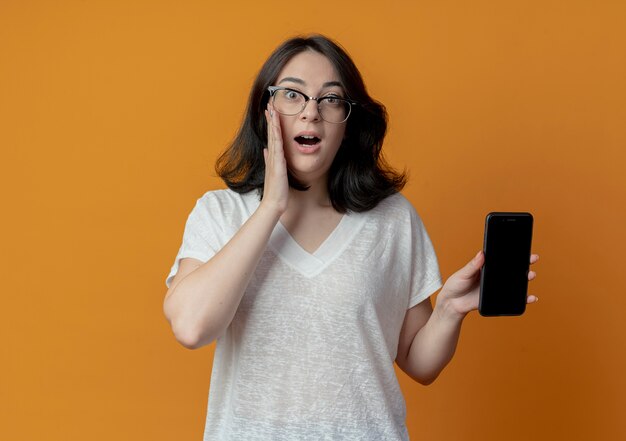 Überraschtes junges hübsches kaukasisches Mädchen, das Brillen hält, die Handy halten und Hand auf Gesicht lokalisiert auf orange Hintergrund mit Kopienraum setzen