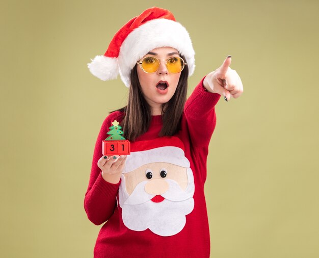 Überraschtes junges hübsches kaukasisches Mädchen mit Weihnachtsmann-Pullover und Stirnband mit Brille, das Weihnachtsbaumspielzeug mit Datum hält und auf die Seite zeigt, isoliert auf olivgrünem Hintergrund
