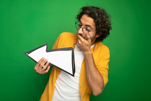 Überraschtes junges hübsches kaukasisches Manntragen der Brille, die Pfeilmarkierung hält, die zur Seite zeigt, die es betrachtet, die Hand auf Mund lokalisiert auf grünem Hintergrund mit Kopienraum hält