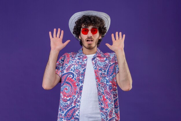 Überraschtes junges hübsches lockiges reisendes Mann, das Sonnenbrille und Hut trägt, die leere Hände auf isolierter lila Wand mit Kopienraum zeigen