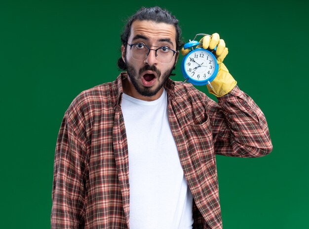 Überraschtes junges hübsches Reinigungspersonal, das T-Shirt und Handschuhe hält, die Wecker halten und auf grüner Wand lokalisiert hören