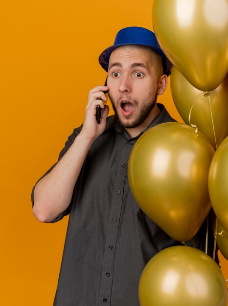 Überraschtes junges hübsches slawisches Partei-Kerl, das Parteihut hält, der Ballons hält, die am Telefon sprechen, das gerade lokalisiert auf orange Hintergrund schaut