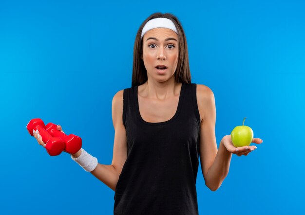 Überraschtes junges hübsches sportliches Mädchen, das Stirnband und Armband hält Hanteln und Apfel lokalisiert auf blauem Raum