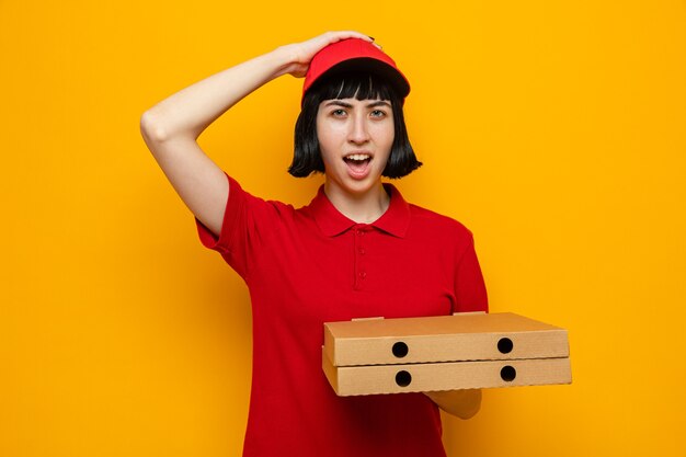 Überraschtes junges kaukasisches Liefermädchen, das Pizzakartons hält und sich die Hand auf den Kopf legt