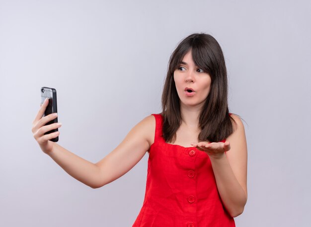 Überraschtes junges kaukasisches Mädchen, das Telefon hält und leere Hand zeigt Kamera auf lokalisiertem weißem Hintergrund betrachtet