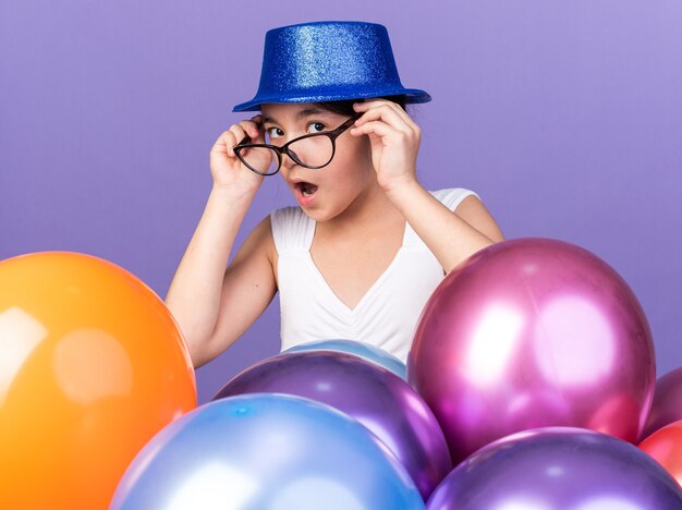 Überraschtes junges kaukasisches Mädchen in optischer Brille mit blauem Partyhut, der mit Heliumballons isoliert auf lila Wand mit Kopierraum steht standing