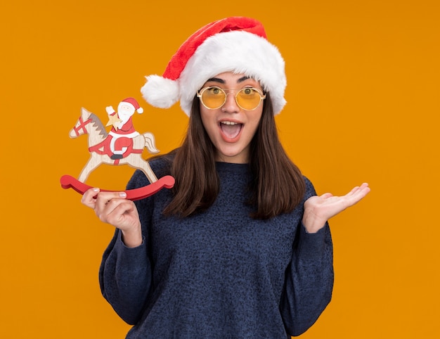 Überraschtes junges kaukasisches Mädchen in Sonnenbrille mit Weihnachtsmütze hält den Weihnachtsmann auf der Schaukelpferddekoration und hält die Hand isoliert auf der orangefarbenen Wand mit Kopierraum offen