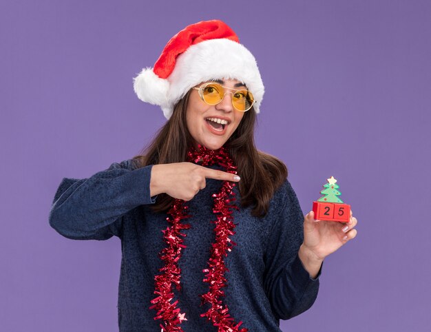 Überraschtes junges kaukasisches Mädchen in Sonnenbrille mit Weihnachtsmütze und Girlande um den Hals hält und zeigt auf Weihnachtsbaumschmuck einzeln auf lila Wand mit Kopierraum