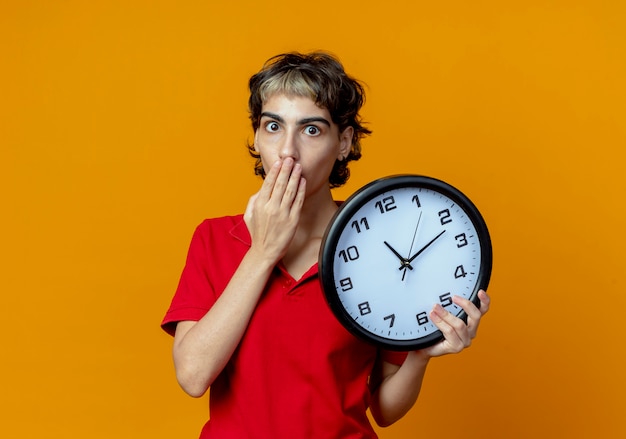 Überraschtes junges kaukasisches Mädchen mit Pixie-Haarschnitt, der Uhr hält und Hand auf Mund lokalisiert auf orange Hintergrund mit Kopienraum setzt