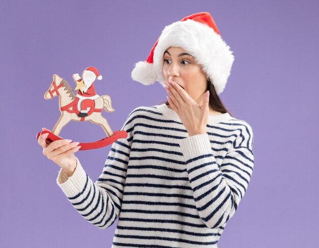 Überraschtes junges kaukasisches Mädchen mit Weihnachtsmütze legt die Hand auf den Mund und sieht den Weihnachtsmann auf Schaukelpferddekoration einzeln auf lila Wand mit Kopienraum an
