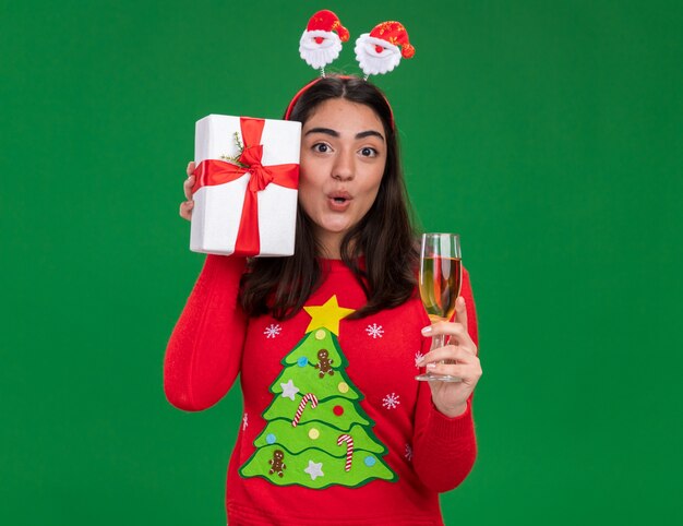 Überraschtes junges kaukasisches Mädchen mit Weihnachtsstirnband hält ein Glas Champagner und eine Weihnachtsgeschenkbox isoliert auf grüner Wand mit Kopierraum