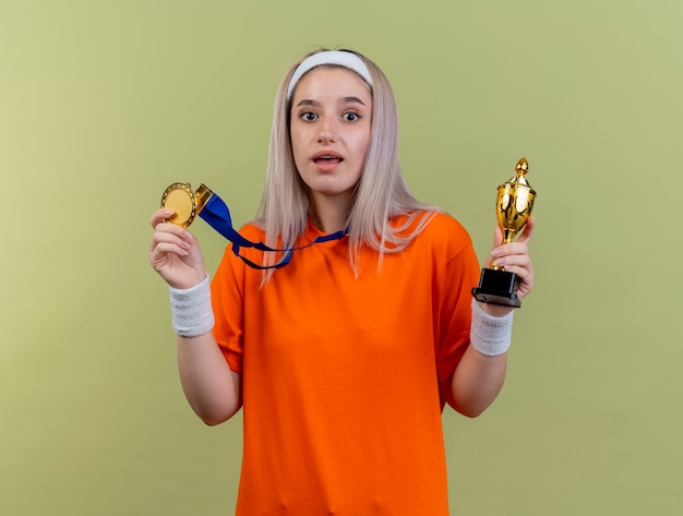 Überraschtes junges kaukasisches sportliches Mädchen mit Hosenträgern, das Stirnband und Armbänder trägt, hält Goldmedaille und Siegerpokal