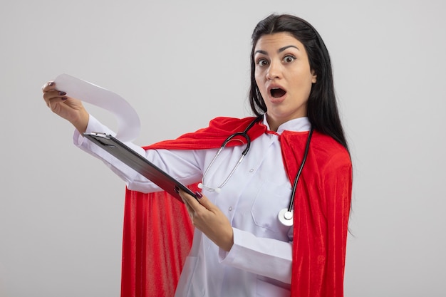 Überraschtes junges kaukasisches Superheldenmädchen, das Stethoskop hält, das Zwischenablage hält, die Kamera lokalisiert auf weißem Hintergrund betrachtet