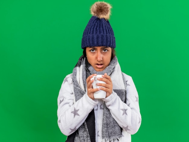 Überraschtes junges krankes Mädchen, das Wintermütze mit Schal hält Tasse Tasse lokalisiert auf grünem Hintergrund trägt