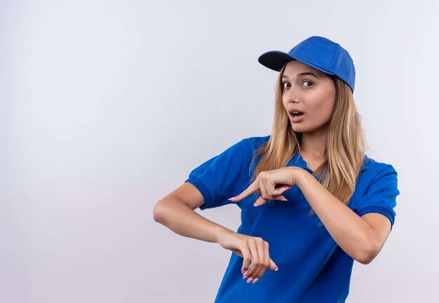 Überraschtes junges Liefermädchen, das blaue Uniform und Kappe trägt, die Handgelenkgeste zeigt, lokalisiert auf weißer Wand mit Kopienraum