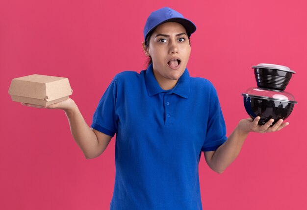 Überraschtes junges Liefermädchen, das Uniform mit Kappe hält, die Lebensmittelbehälter hält, die Hände lokalisiert auf rosa Wand spreizen