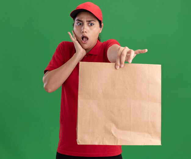 Überraschtes junges Liefermädchen, das Uniform und Kappe trägt, die Papiernahrungsmittelpaket an der Kamera hält, die Hand auf Wange lokalisiert auf grüner Wand setzt