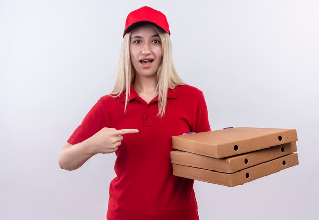 Überraschtes junges Mädchen der Lieferung, das rotes T-Shirt und Kappe in Zahnspange trägt, zeigt auf Pizzakiste auf ihrer Hand auf lokalisiertem weißem Hintergrund