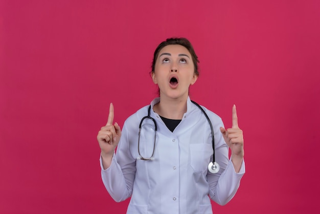 Überraschtes junges Mädchen des Arztes, das medizinisches Kleid und Stethoskop trägt, zeigt oben auf isoliertem rotem Hintergrund