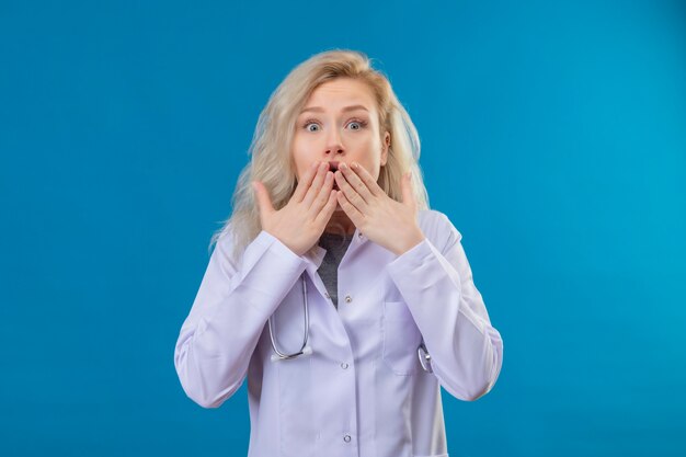 Überraschtes junges Mädchen des Arztes, das Stethoskop im medizinischen Kleid bedeckte, bedeckte Mund auf blauem Hintergrund