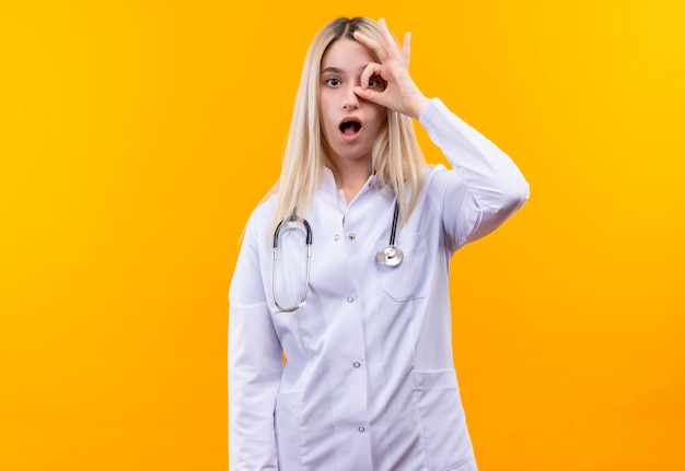 Überraschtes junges Mädchen des Arztes, das Stethoskop im medizinischen Kleid trägt, das Blickgeste auf lokalisiertem gelbem Hintergrund zeigt