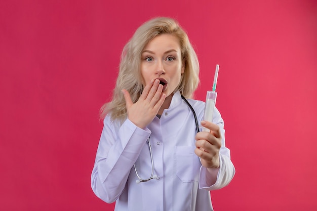 Überraschtes junges Mädchen des Arztes, das Stethoskop im medizinischen Kleid trägt Spritzen bedeckte Mund auf rotem Hintergrund