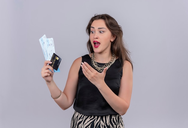 Überraschtes junges Mädchen des Reisenden, das schwarzes Unterhemd hält, das Tickets und Kreditkarte auf weißem Hintergrund hält