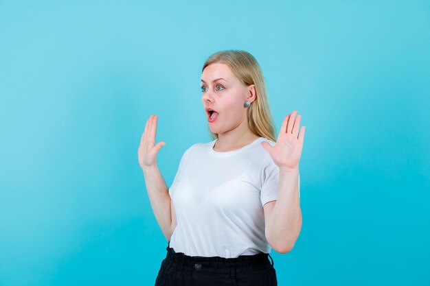 Überraschtes junges Mädchen schaut weg, indem sie ihre Handvoll auf blauem Hintergrund erhebt