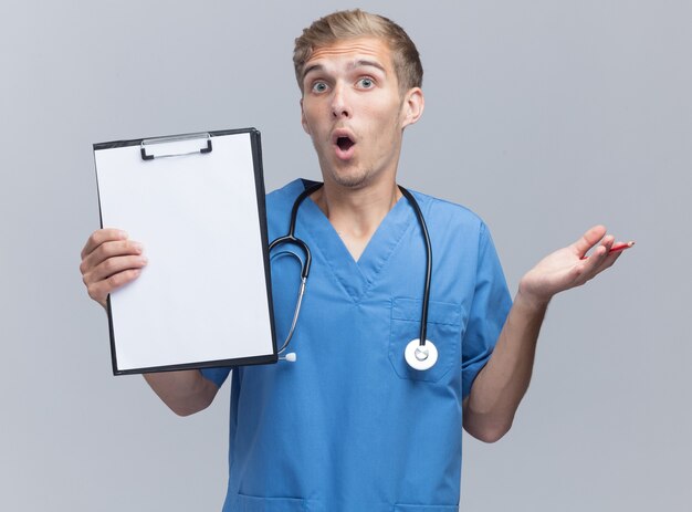 Überraschtes junges männliches Doktor tragen Arztuniform mit Stethoskop, das Klemmbrett hält und Hand lokalisiert auf weißer Wand spreizt