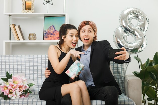 Überraschtes junges Paar am glücklichen Frauentag, das Geschenk hält, macht ein Selfie, das auf dem Sofa im Wohnzimmer sitzt