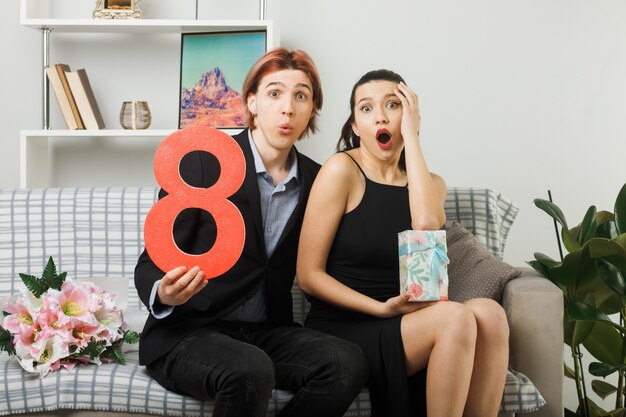 Überraschtes junges Paar am glücklichen Frauentag mit Nummer acht mit anwesendem Mädchen, das die Hand auf die Wange legt, die auf dem Sofa im Wohnzimmer sitzt