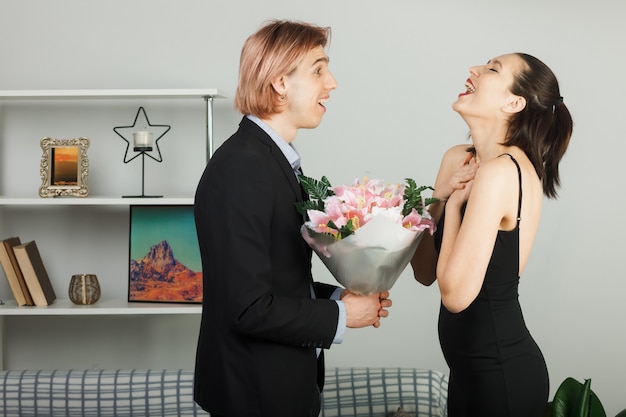 Überraschtes junges Paar umarmte sich am Valentinstag mit Blumenstraußmädchen, die im Wohnzimmer stehen