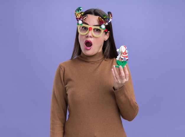 Überraschtes junges schönes Mädchen, das braunen Pullover mit Weihnachtsbrille hält Spielzeug lokalisiert auf blauem Hintergrund trägt