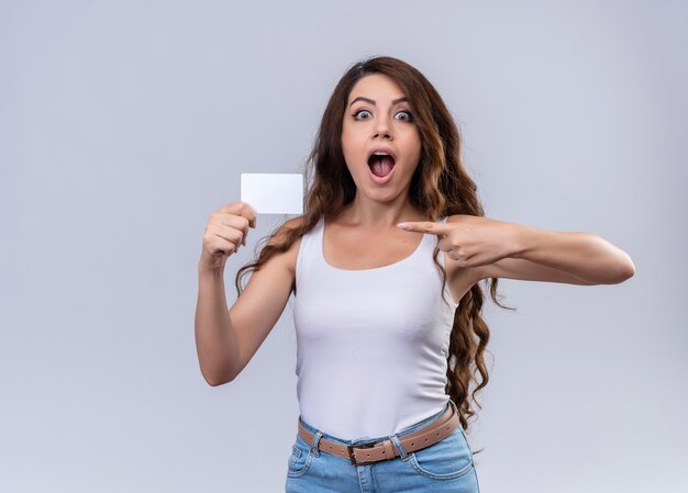 Überraschtes junges schönes Mädchen, das Kreditkarte hält und auf lokalisierte weiße Wand mit Kopienraum zeigt