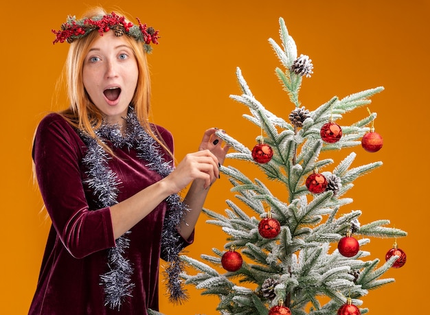 Überraschtes junges schönes Mädchen, das nahe Weihnachtsbaum steht, der rotes Kleid und Kranz mit Girlande auf Hals lokalisiert auf orange Hintergrund trägt