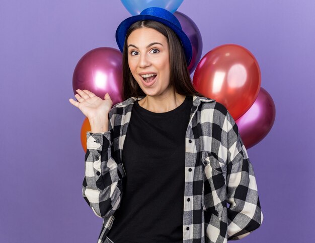 Überraschtes junges schönes Mädchen mit Partyhut, das vor Ballons steht