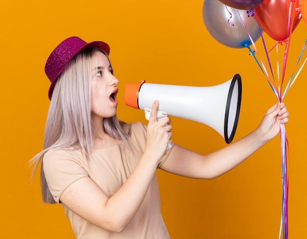 Überraschtes junges schönes Mädchen mit Partyhut mit Luftballons spricht über Lautsprecher