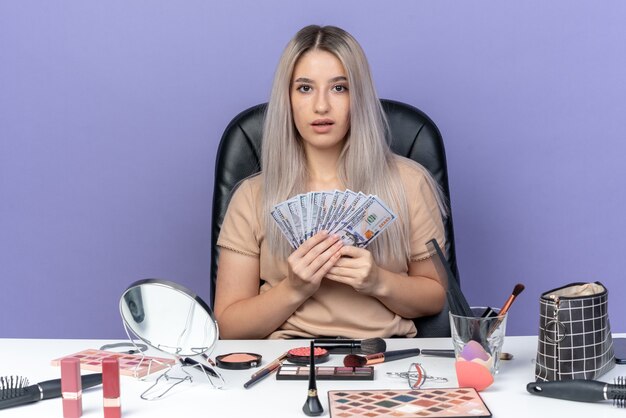 Überraschtes junges schönes Mädchen sitzt am Tisch mit Make-up-Tools, die Bargeld auf blauem Hintergrund halten