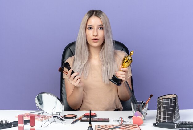 Überraschtes junges schönes Mädchen sitzt am Tisch mit Make-up-Tools, die Gewinnerbecher mit Make-up-Pinsel auf blauem Hintergrund halten