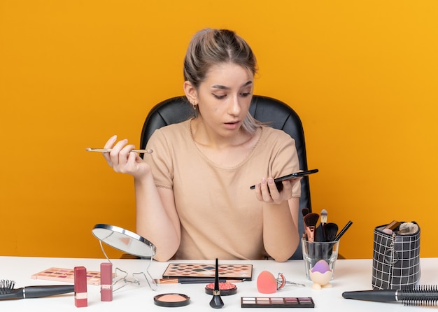 Überraschtes junges, schönes Mädchen sitzt am Tisch mit Make-up-Tools, die Make-up-Pinsel halten und das Telefon in der Hand einzeln auf orangefarbenem Hintergrund betrachten