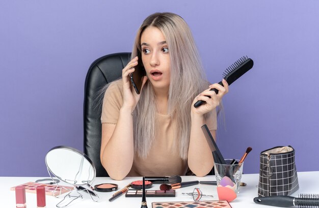 Überraschtes junges schönes Mädchen sitzt am Tisch mit Make-up-Tools spricht am Telefon mit Kamm isoliert auf blauem Hintergrund